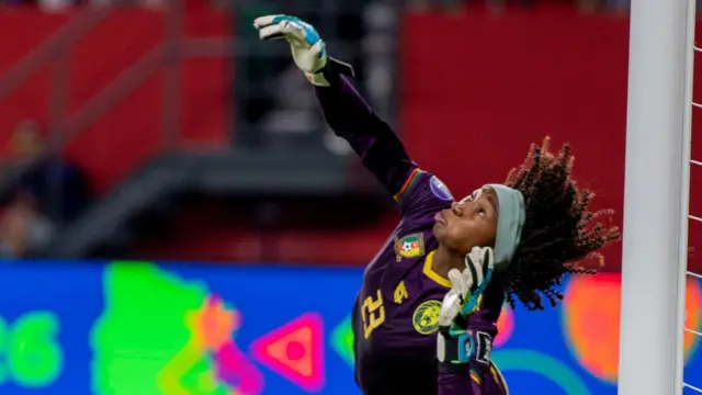 La gardienne camerounaise Michaely Bihina, vêtue d'une tenue entièrement violette et d'un large bandeau bleu, cambre le dos en levant le bras droit pour repousser un penalty.