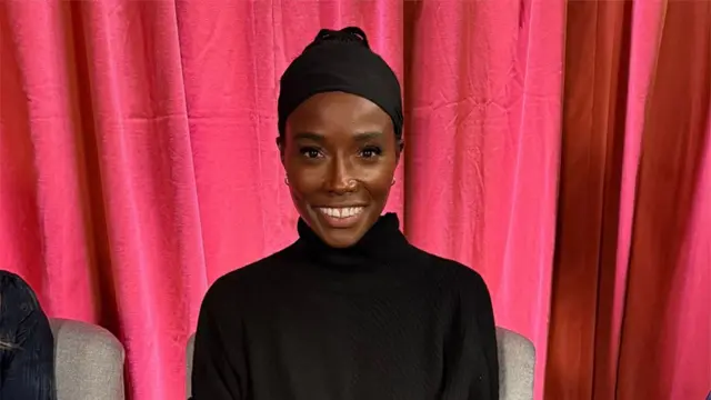 赤いカーテンを背に、黒い服と黒いヘッドバンドを付けたアフリカ系女性がカメラに笑顔を向けている