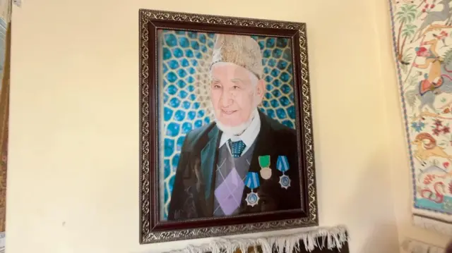 حاجی محمدعوض بادغیسی در این تصویر با لبخند به کمره نگاه می‌کند. کلاه پوست بر سر دارد و کت‌وشلوار همراه با نکتایی پوشیده است. چندین مدال و نشان افتخار که به پاس فعالیت‌ها و دستاوردهایش دریافت کرده، بر سینه او دیده می‌شود.
