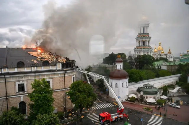 Велика історична будівля з пошкодженим дахом, який горить, а над нею піднімається густий чорний дим. Рятувальники на червоній машині використовують високу драбину, щоб гасити полум’я на даху. Поруч – церква з пошкодженим куполом.