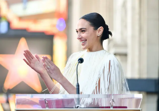 Gal Gadot kürsüde beyaz elbisesiyle alkış çalıyor. 