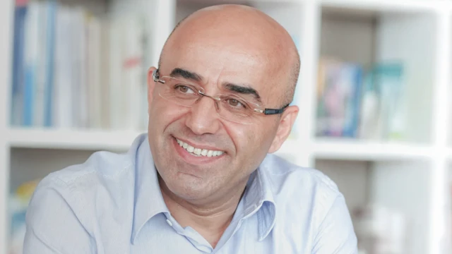 Necati Özkan gülerek poz veriyor. Fotoğrafta gözlüklü ve kravatsız açık mavi bir gömlek giyiyor.