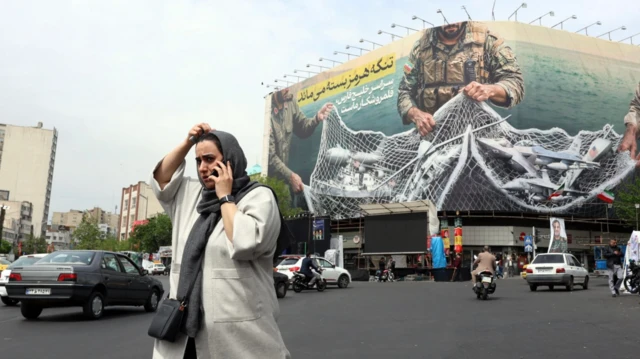Bir kadın büyük bir caddenin ortasından telefonla konuşarak geçiyor. Arkada dev bir İran'ın savaşını destekleyen poster var.