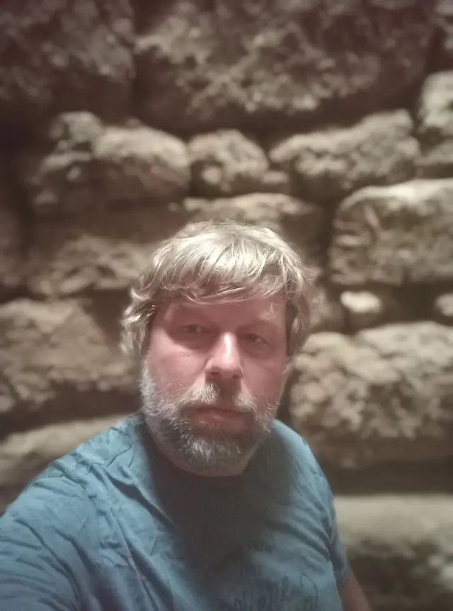 Археолог Бутягин с бородой и в футболке на фоне камней