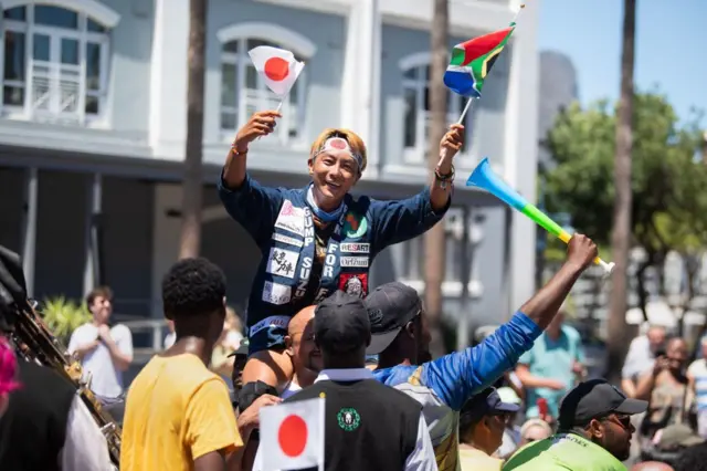 A man waves the flags of Kenya and South Africa on Cape Town's waterfront. People hold him up in the air and celebrate with him.