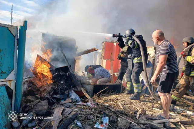 Рятувальники гасять пожежу серед зруйнованих будівель і палаючих уламків, спрямовуючи струмінь води на вогонь. Поряд стоїть цивільна людина, а ще одна людинa схилилася біля землі; довкола — дим, вогонь і розкидані рештки.
