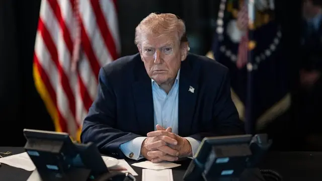 Donald Trump. Viste un traje oscuro con una camisa clara. Está sentado en un escritorio sobre el que apoya sus dos manos entrelazadas mientras mira al frente. Al fondo, a la izquierda hay una bandera de EE.UU.