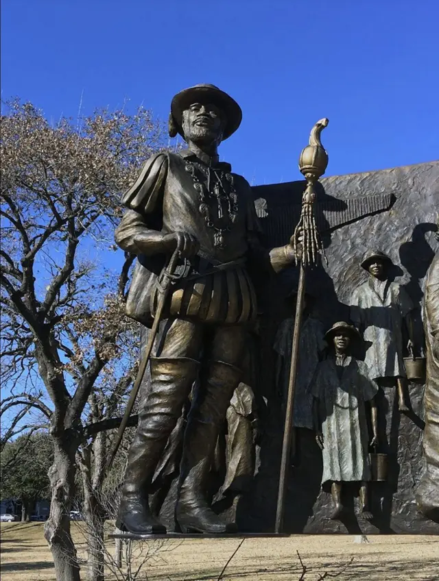 Monumento de bronce con una figura histórica sosteniendo un bastón ceremonial, frente a un relieve escultórico y bajo un cielo despejado.