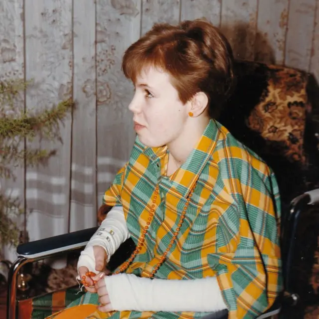 1982 tarihli renkli bir fotoğrafta, 11 yaşındaki Nina bir oturma odasında tekerlekli sandalyede otururken görülüyor. Fotoğraftaki kişi, açık renkli uzun kollu bir üstün üzerine yeşil ve sarı kareli bir kıyafet giymiş ve elinde turuncu boncuklardan oluşan bir kolye tutuyor. Arka planda dantel perdeler bir pencereyi örtüyor ve tekerlekli sandalyenin yanında desenli bir koltuğun bir kısmı görünüyor. 