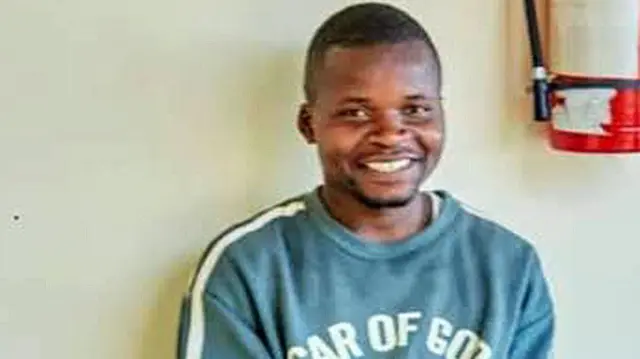 Fọto yii ṣafihan Shedrack Chaula lati ori de ejika rẹ, o n rẹrin-in. Aṣọ alawọ ewe lo wọ, irinṣẹ ti wọn fi n pa ina si wa lara ogiri ibi to duro si.