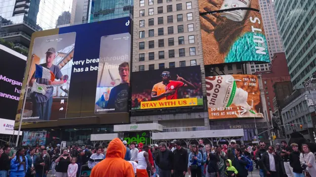 A billboard bearing a picture of Victor Osimhen and a message saying 'stay wit us' dey seen among oda electronic advertising hoardings above a crowded street in New York