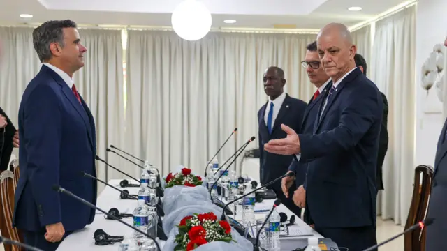 Uzun bir masa boyunca plastik su şişeleri ve ince mikrofonlarla birlikte takım elbiseli erkekler ayakta duruyor.