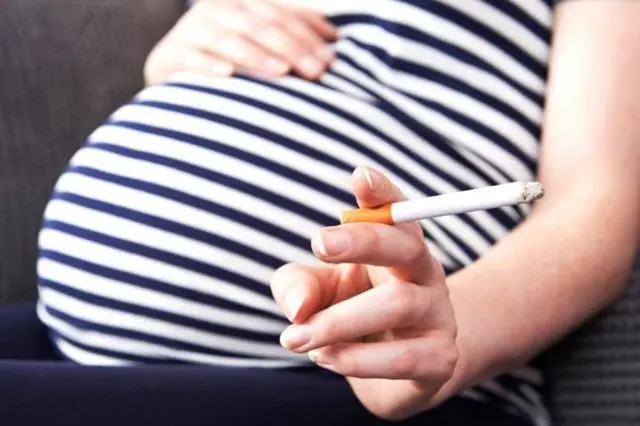 Pregnant woman in blue and white striped top with right hand resting on baby bump and left hand holding a lit cigarette.
