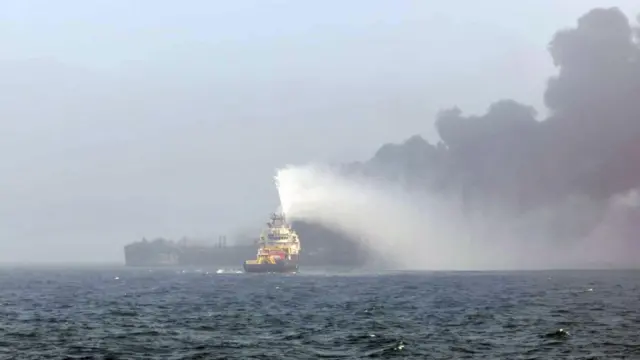 Foto de um navio com esguicho de água em direção a uma nuvem de fumaça preta em alto-mar. 