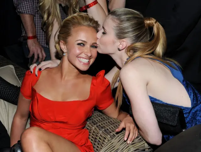 Hayden Panettiere, con un vestido rojo de largo medio y el pelo recogido, sonríe mientras su compañera de reparto, Emma Roberts, le da un beso en la mejilla en la fiesta posterior al estreno de Scream 4.