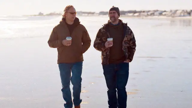 গ্রেগ স্কয়ার এবং পিট ম্যানিং সমুদ্র সৈকতে টেকওয়ে পানীয় হাতে হাঁটছেন। তারা দুজনেই নীল প্যান্ট এবং বাদামী জ্যাকেট পরে আছেন।