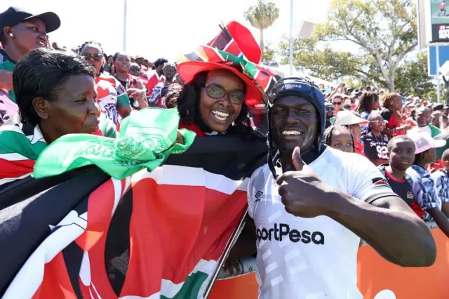 A man poses with his thumbs up. Next to him a woman waves the Kenyan flag.
