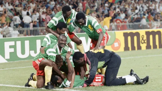 Les joueurs de champ extérieur camerounais vêtus de chemises vertes, de shorts rouges et de chaussettes jaunes, ainsi que le gardien de but vêtu principalement de noir, y compris son pantalon de survêtement, célèbrent un but lors de la Coupe du monde 1990 en s'entassant sur le dessus d'Eugene Ekeke, allongé sur le dos alors que six joueurs se rassemblent autour de lui pour le féliciter. Des panneaux publicitaires peuvent être vus en arrière-plan, avec un stand bondé de fans au loin. Un joueur, Roger Milla, lève les yeux du peloton et sourit à la caméra