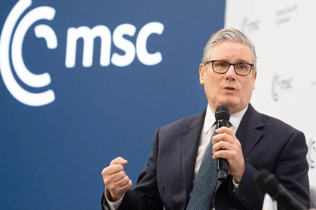 Keir Starmer veste um terno escuro, gravata azul e está segurando um microfone com sua mão esquerda na Conferência de Segurança de Munique