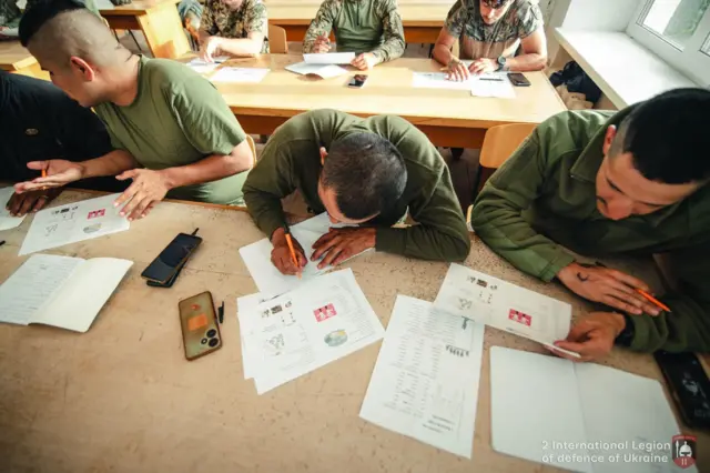 Ряды парты, за передней сидят трое мужчин в одежде военного типа, за партой позади их еще трое. Перед учениками лежат цветные распечатки с заданиями.
