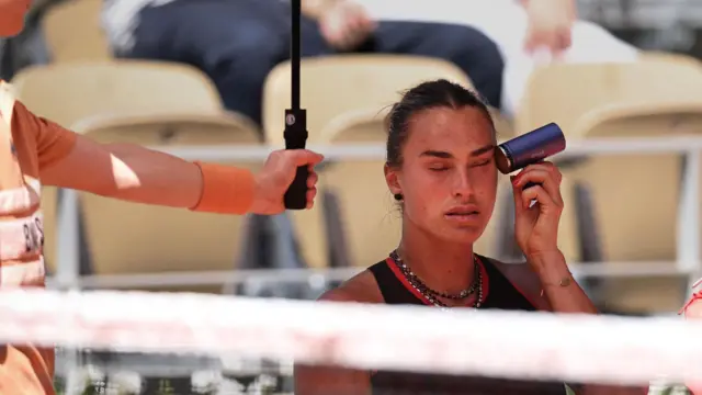 Aryna Sabalenka chłodzi się na korcie. Trzyma przy skroni mały przenośny wentylator 