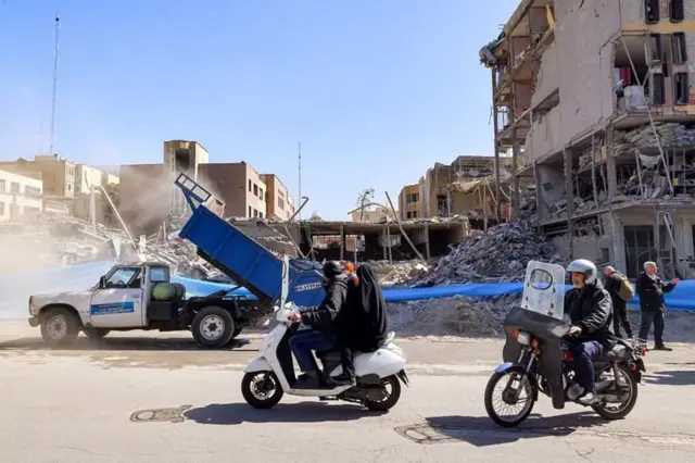 मोटरसाइकिल पर सवार लोग तेहरान के फिरदौसी स्क्वायर के पास ध्वस्त हुई इमारतों से गुजरते हैं. यहां कल हमला हुआ था 