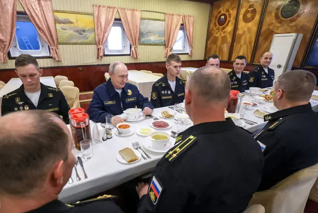 Путин сидит за длинным обеденным столом, накрытым белой скатертью, вместе с офицерами «Варяга». На столе различные закуски и тарелки с супом