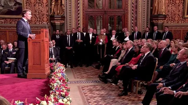 大広間の演壇を横から撮った写真。左手の壇上にマクロン氏がたち、右側のフロアに椅子が並べられ、議員らが座っている。奥の扉の前にも人々が立っている