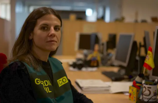 Cristina with a police vest that says Guardia Civil, sitting at her desk next to a row of computers, with a small Spanish flag next to a monitor