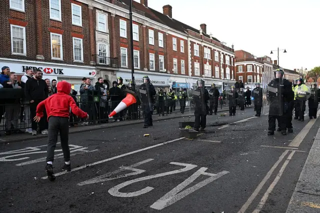 A protester throws a traffic cone on a road in Epsom as riot police walk towards the protester with shields.