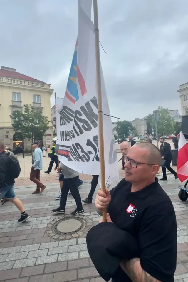 Чоловік у темній футболці та окулярах тримає високий банер із перекресленим тризубом на дерев’яному держаку. На грудях видно два невеликі символи: червоно-білий значок із польським орлом та білий кельтський хрест (вертикальний хрест, обведений колом). Навколо йдуть учасники зібрання, поруч видно польський прапор, історичні будівлі та перехожих.