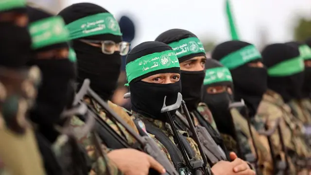 Fila de combatentes do Hamas, de uniforme e com espingardas em punho. Seu rosto é coberto por uma balaclava preta e, na testa, levam uma faixa verde característica do grupo