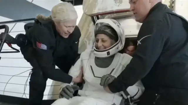 Suni Williams sai da cápsula sorrindo, vestindo um traje espacial branco e capacete, auxiliada por dois assistentes vestidos de preto.