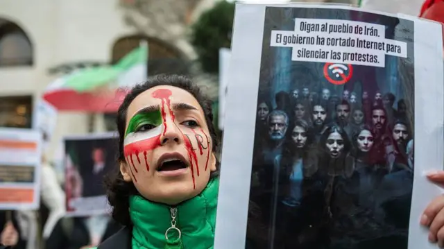 مادرید، اسپانیا – ۲۲ ژانویهٔ ۲۰۲۶:
زنی با صورت نقاشیشده به رنگهای پرچم ایران و یک زخم گلولهٔ نمادین بر سر، در جریان اعتراضی مقابل پارلمان اسپانیا دیده میشود؛ او پلاکاردی در دست دارد که علیه قطع اینترنت در ایران است.