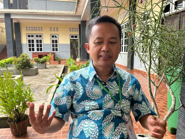 Seorang pria sedang menggunakan baju batik sedang menjelaskan sesuatu.