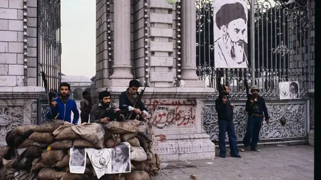 Des soldats et des hommes armés montent la garde devant un bâtiment à Téhéran, avec une affiche de Khomeini accrochée en arrière-plan.