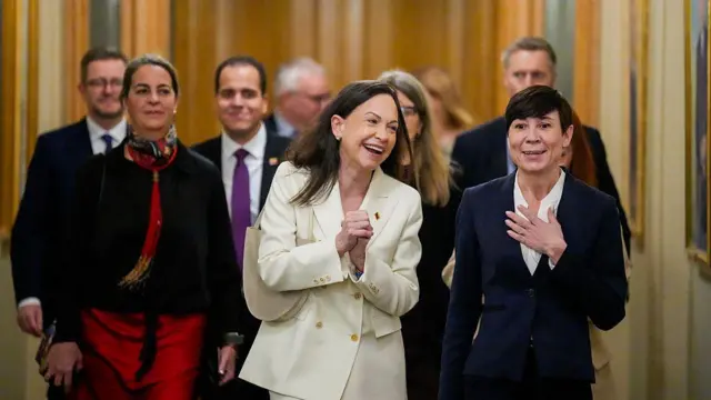 María Corina Machado camina hacia una conferencia de prensa en oslo, noruega, este jueves. Viste un chaleco blanco, sonríe mientras camina con un grupo de personas. Lleva el cabello negro hasta los hombros y tiene sus manos juntas. 
