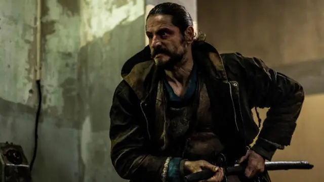 Мушкарац са црном везаном косом и брадом, у тамној јакни гледа у страну и држи се за мач око појаса