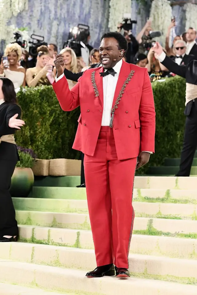 Broadway oyuncusu Joshua Henry, gümüş yakalı işlemeli kırmızı kruvaze bir smokin giyiyor. Merdivenlerde durup basın mensupları için performans sergilerken şarkı söylüyor.