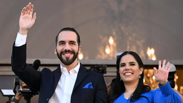 El Salvador Devlet Başkanı Nayib Bukele, eşi Gabriela Roberta Rodríguez de Bukele'nin yanında gülümseyerek el sallıyor. Arka planda büyük bir avize asılı duruyor ve Nayib Bukele'nin önünde mikrofon ayakları yer alıyor. Kendisi lacivert bir takım elbise ve beyaz gömlek giyerken, Gabriela mavi bir üst giyiyor.
