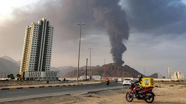 دراجة نارية متوقفة على جانب الطريق وفي الخلفية تلال ترابية يتصاعد من خلفها دخان أسود