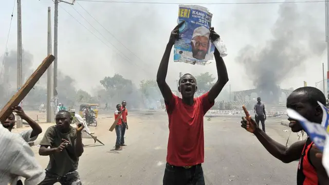 De jeunes manifestants sont en action dans les rues de Kano - l'un d'eux tient une photo de Buhari en l'air, deux autres brandissent des morceaux de bois. De la fumée grise s'échappe à l'arrière-plan.