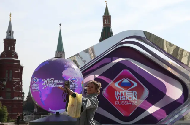 Uma mulher tira uma selfie na Praça Vermelha, em Moscou. Atrás dela, há um grande balão decorativo e um telão com as palavras INTERVISION RUSSIA'25 e o slogan “Música no coração do seu país”.
