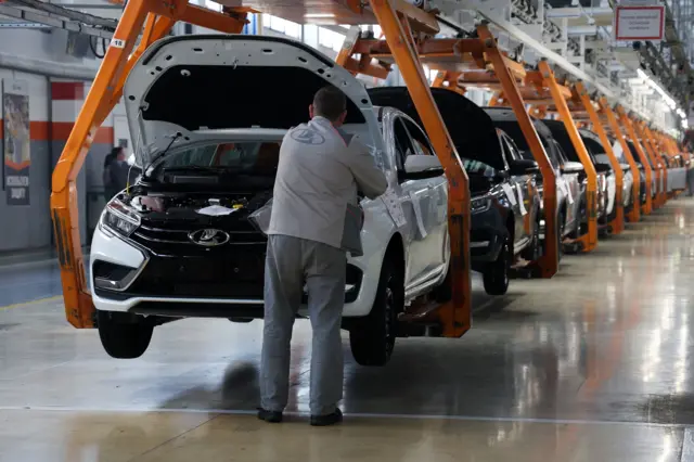 Цех автомобильного завода. В цехе висят автомобили с поднятыми капотами. У одного автомобиля на переднем плане стоит человек в рабочей одежде с логотипом в виде ладьи.