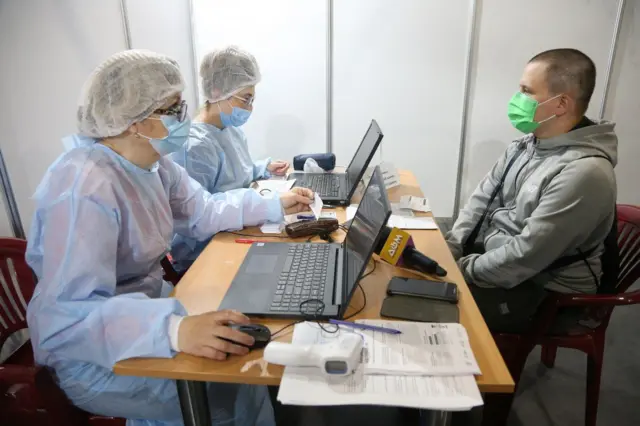 Чоловік у масці сидить за столом перед двома медсестрами