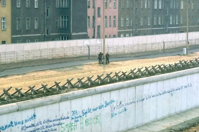 Berlin Duvarı'nın tel örgüleri ve duvar arasında devriye gezen askerlerin uzaktan çekilmiş bir fotoğrafı