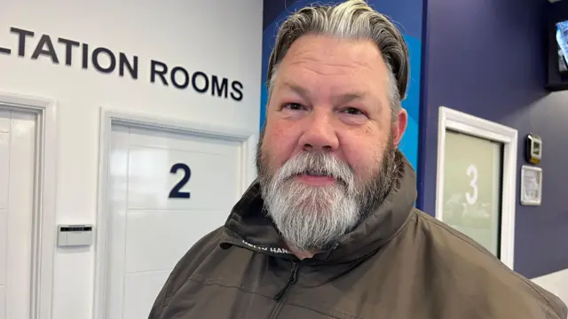 A man with a grey beard and hair looks at the camera. He wears an olive green rain jacket and is standing in front of a sign that reads "CONSULTATION ROOMS" in a pharmacy. 