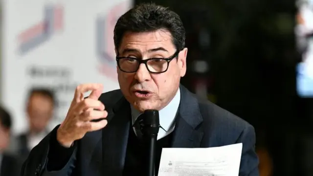 Philippe Aghion falando ao microfone. Ele usa terno e um óculos de grau