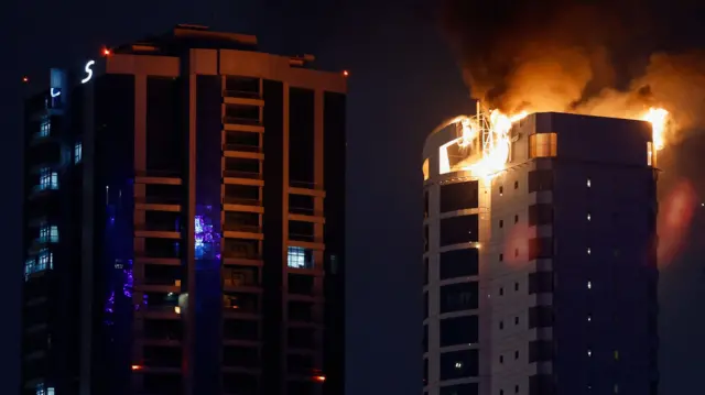 Flames engulf the top of a tower block at night while smoke billows into the night sky. A tall building next to it appears not to have been damaged