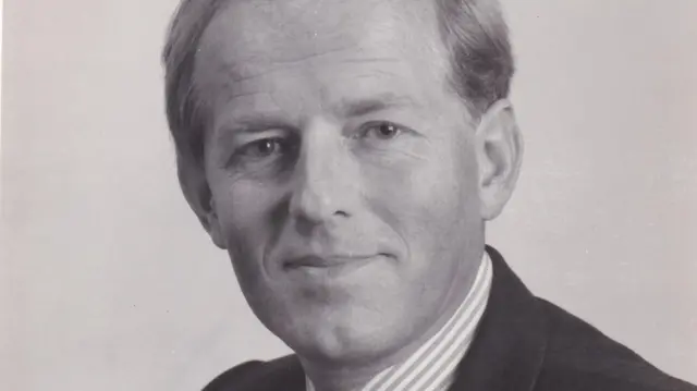 Retrato profesional en blanco y negro de John Smyth, quien viste una camisa de cuello a rayas y un traje oscuro. Sonríe a la cámara y parece amigable.